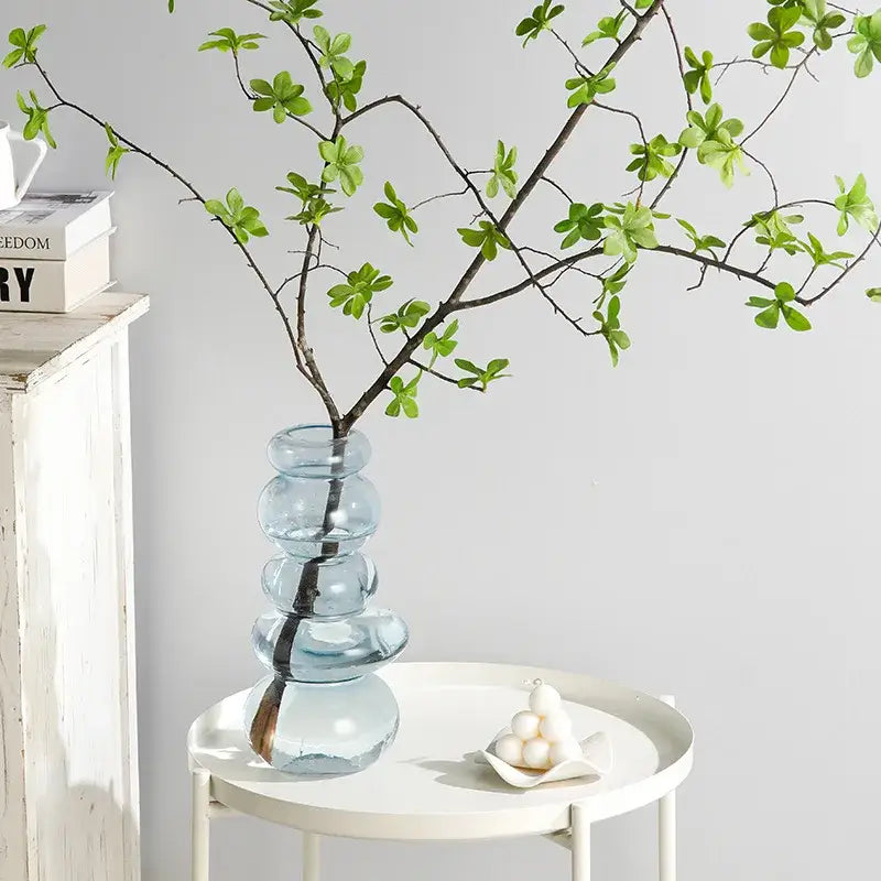 A translucent light blue glass pebble stack floral vase with rounded segments holding a branch of green leaves