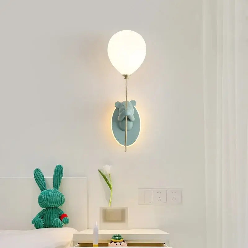A soft white wall lamp shaped like a balloon with a teal decorbites cartoon bear holding the stem on an oval base
