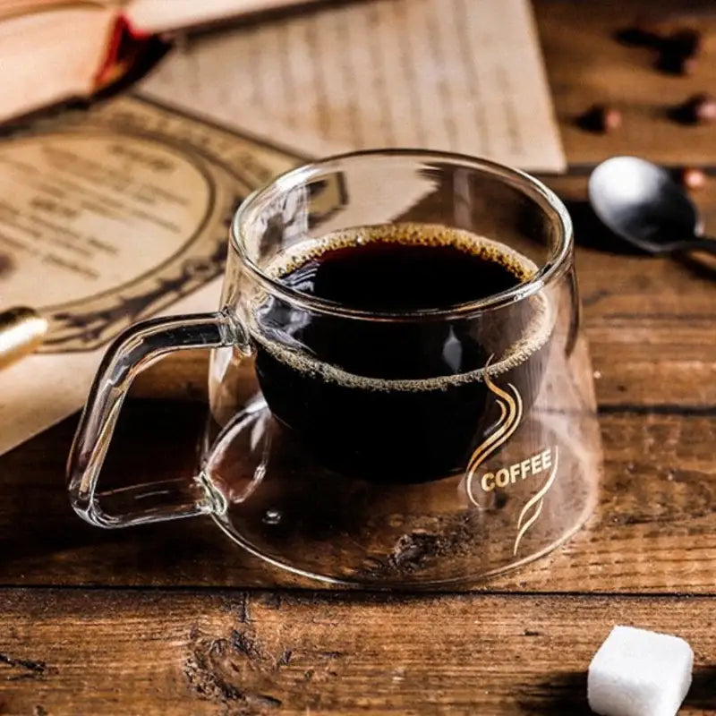 Clear double-walled glass coffee mug with gold coffee logo and swirling design filled with dark espresso decorbites glass double