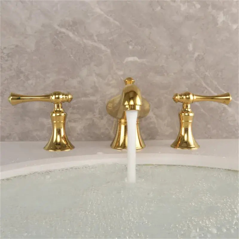 Gold widespread bathroom faucet with three gold finished lever handles and waterfall design set against marble backsplash