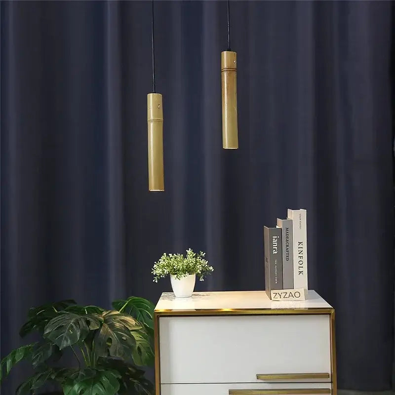 Two sleek cylindrical gold bamboo tube pendant lights with adjustable black cords against a dark blue curtain