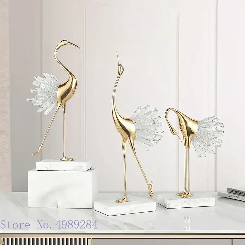 Three elegant gold-colored golden bird agate crane statues with crystal feathers on white bird agate marble bases for home decor