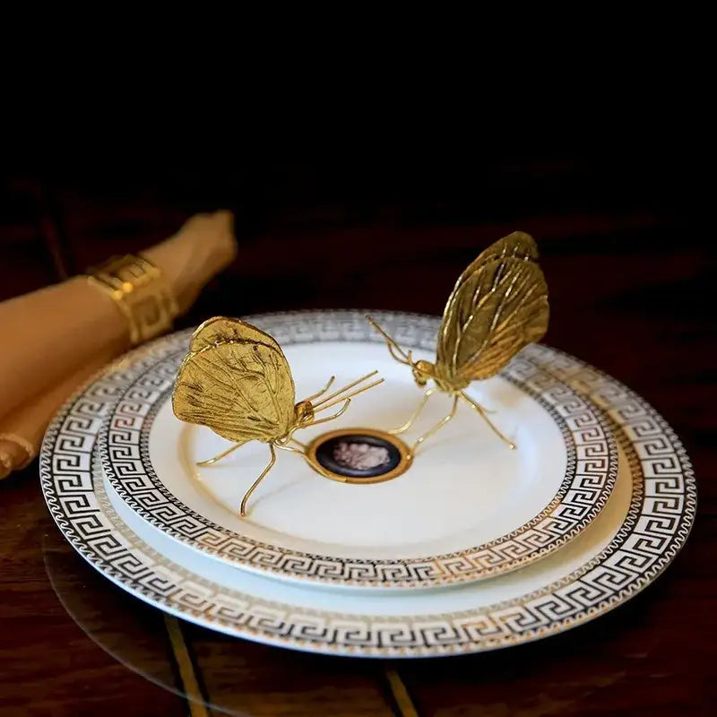 Golden butterfly ant miniature metal figurines with intricate wing detailing on a white plate with black greek key border