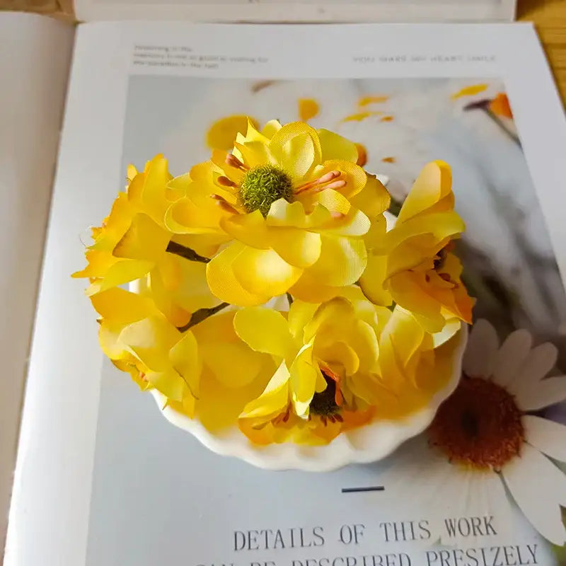 A cluster of vibrant yellow artificial flowers with berry stamen and green plant foliage in a white ceramic bowl for decorbites decor