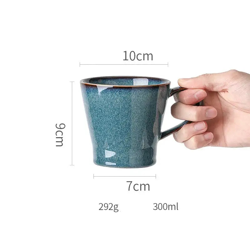 A hand holds a beautiful blue ceramic mug with speckled teal glaze and dark brown rim, complementing dinnerware dish