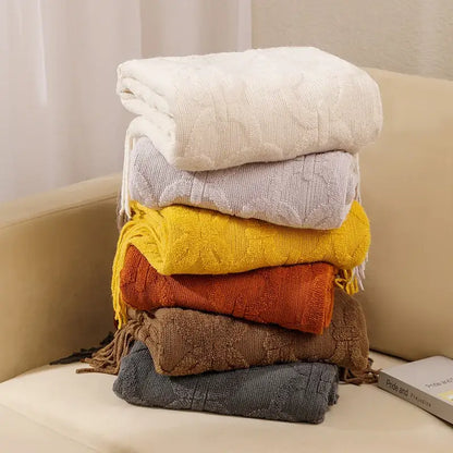 Stack of six decorbites knit blankets with cable knit patterns and fringed edges in cream, gray, yellow, orange, brown, and dark gray for sofa decor