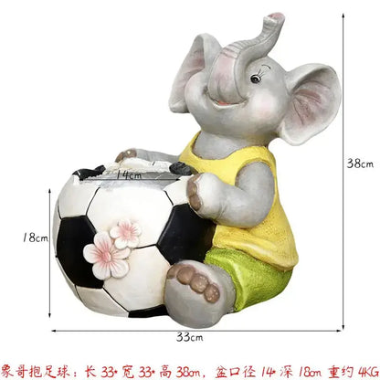 A gray ceramic elephant garden decoration wearing a yellow tank top and green shorts with a floral soccer ball