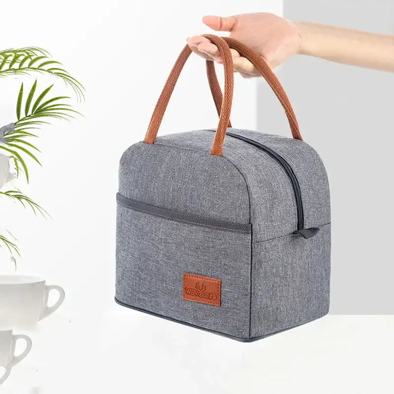 Gray textured insulated lunch bag with brown woven handles and leather patch for thermal food storage