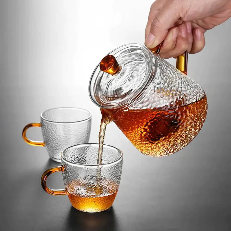 Hammered clear borosilicate glass teapot with amber handle pouring golden tea into matching glass mugs