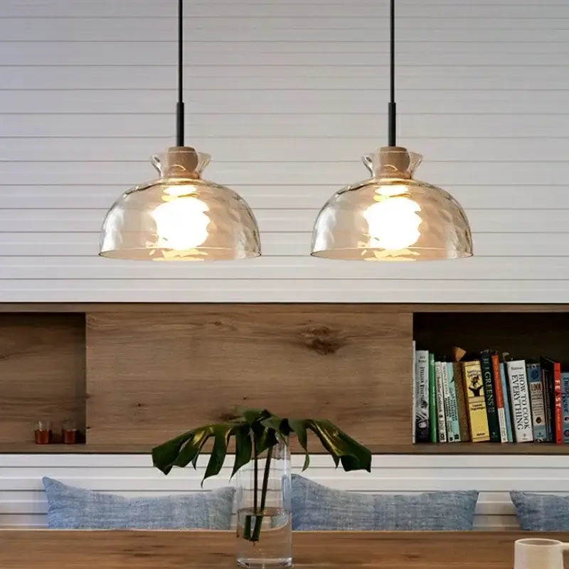 Two warm-toned hammered clear glass pendant lights with black cords hang above a wooden table in a kitchen setting