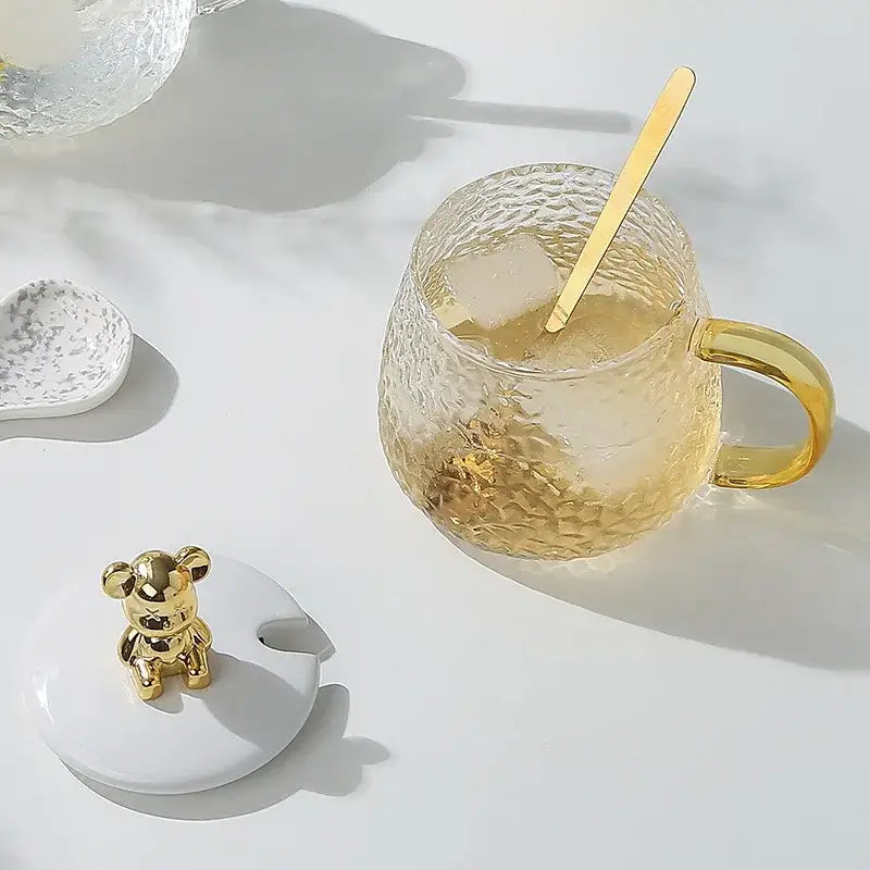 Gold-toned hammered glass water cup with textured gold handle spoon and white ceramic lid with golden bear figurine