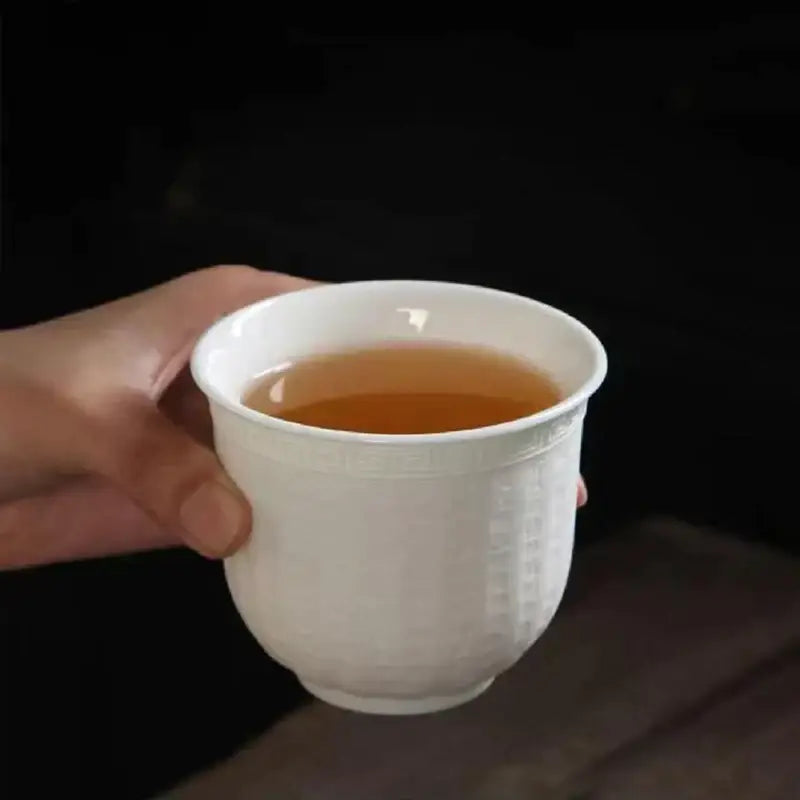 White ceramic tea cup with embossed patterns holding amber tea from hand carved translucent white jade tea cup set
