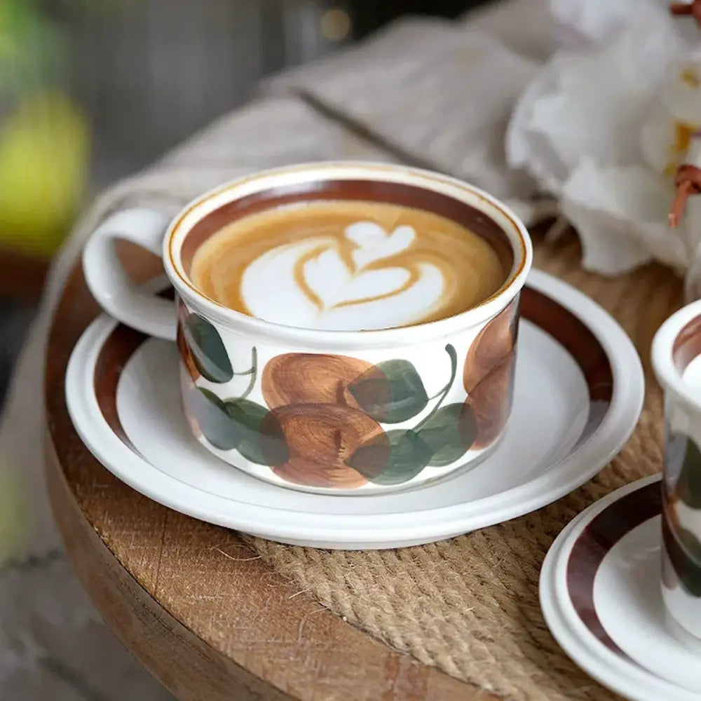 White ceramic espresso cup with brown and green hand-drawn leaf design and heart-shaped latte art on top