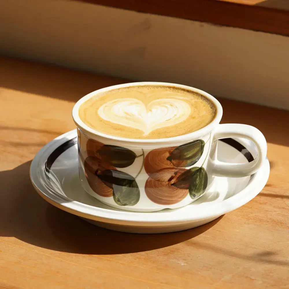 White ceramic espresso cup with hand-drawn brown and green leaf patterns filled with heart-shaped latte art