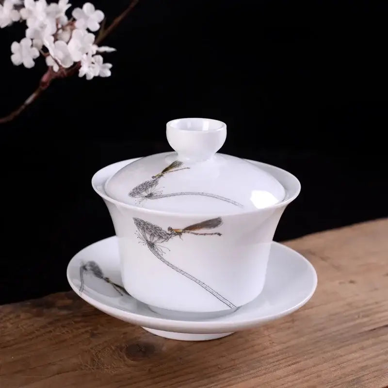 White porcelain gaiwan hand painted with black and gold dragonfly designs resting on a matching saucer decorbites blue