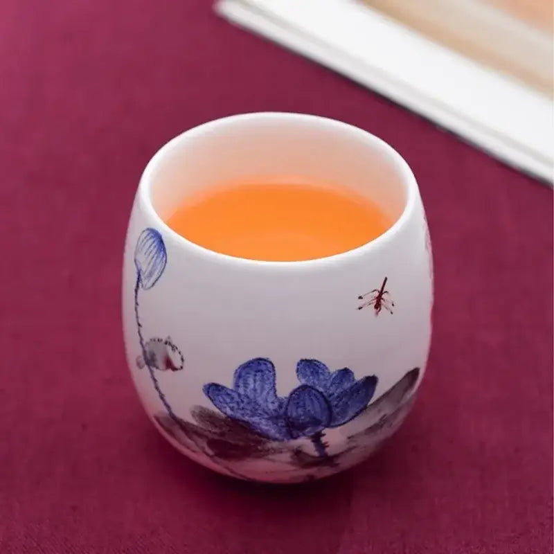 White porcelain kung fu tea cup with hand-painted blue lotus and dragonfly motifs filled with orange liquid decorbites ceramic kung