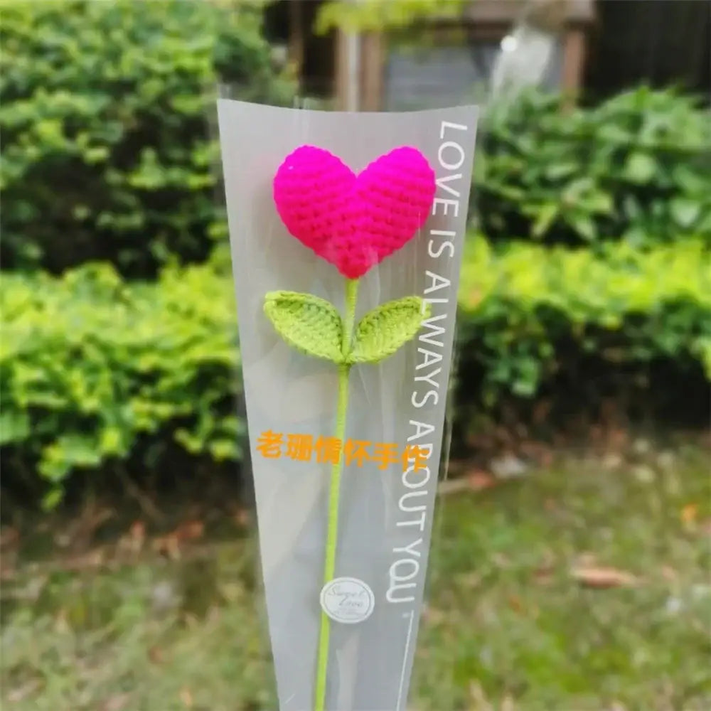 A vibrant pink yarn crochet hand woven love heart flower with green leaves in a clear plastic sleeve for bedroom decor