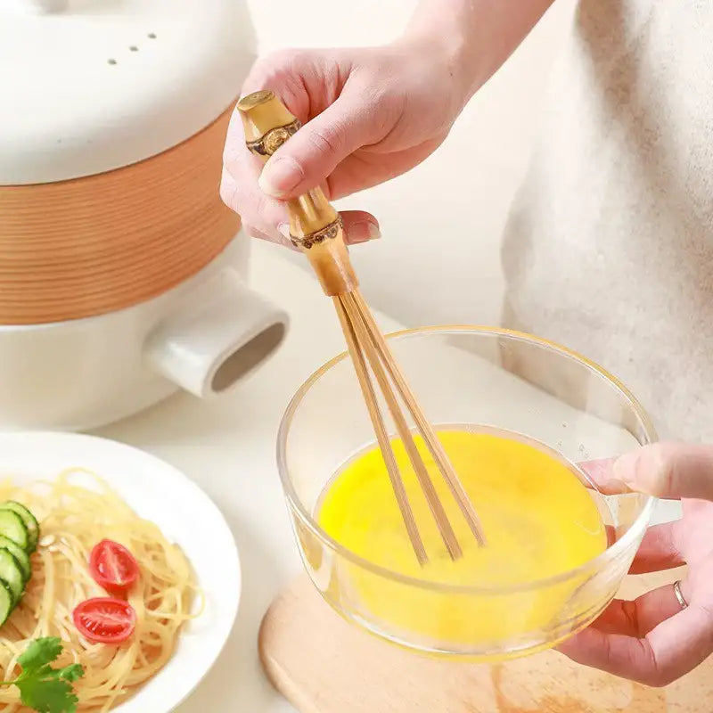 Handmade golden bamboo flour mixer egg beater with bamboo handle beating eggs in a clear glass bowl