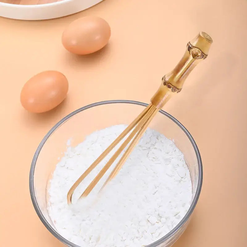 Handmade golden bamboo egg beater with flour mixer blender resting in a glass bowl of white flour