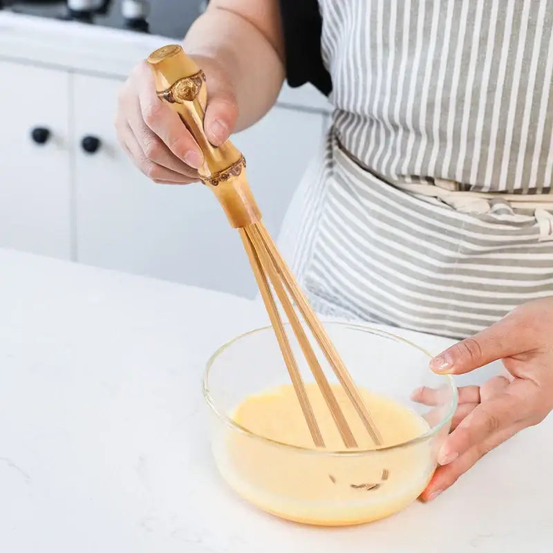 Handmade golden bamboo egg beater with ornate detailing mixing yellow liquid in a clear glass bowl for flour mixer use