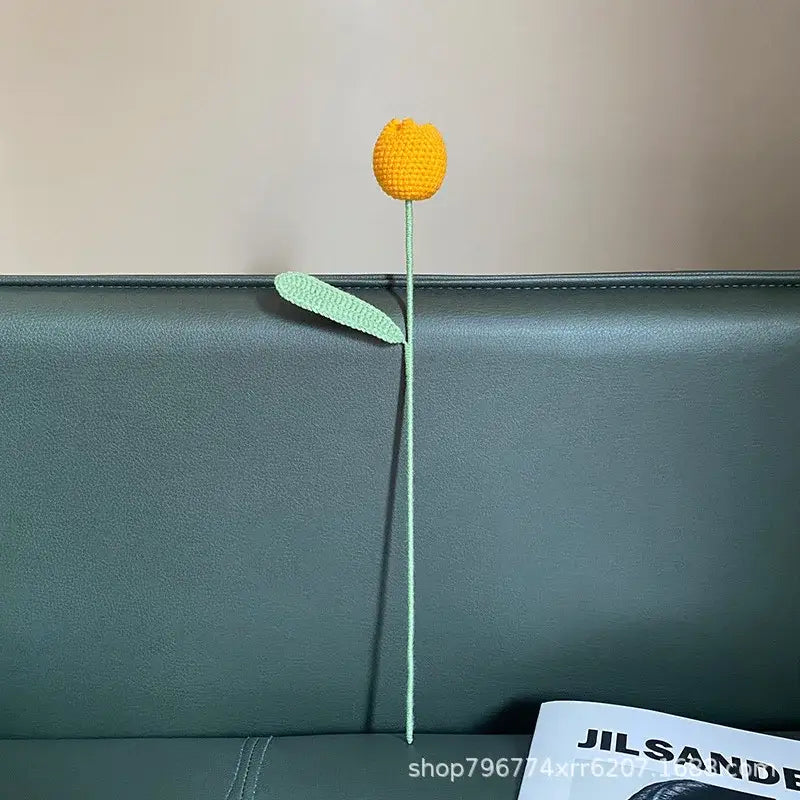 A decorbites handmade knitted yellow crocheted flower with mint green stem resting on a dark leather couch