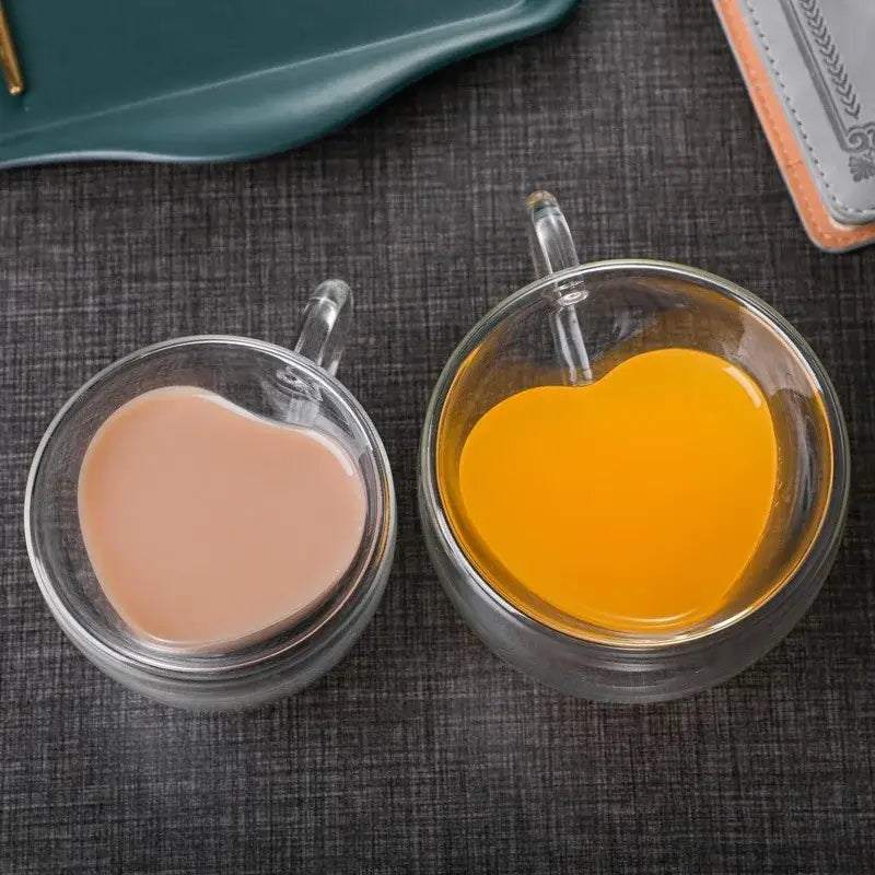 Two clear double walled heart love glass coffee mugs filled with pink drink and orange juice in a mug cup set