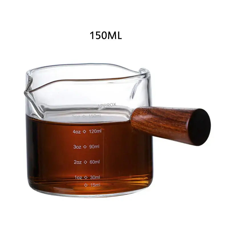 A heat resistant glass espresso milk jug with wooden handle filled with dark liquid marked up to 150ml capacity