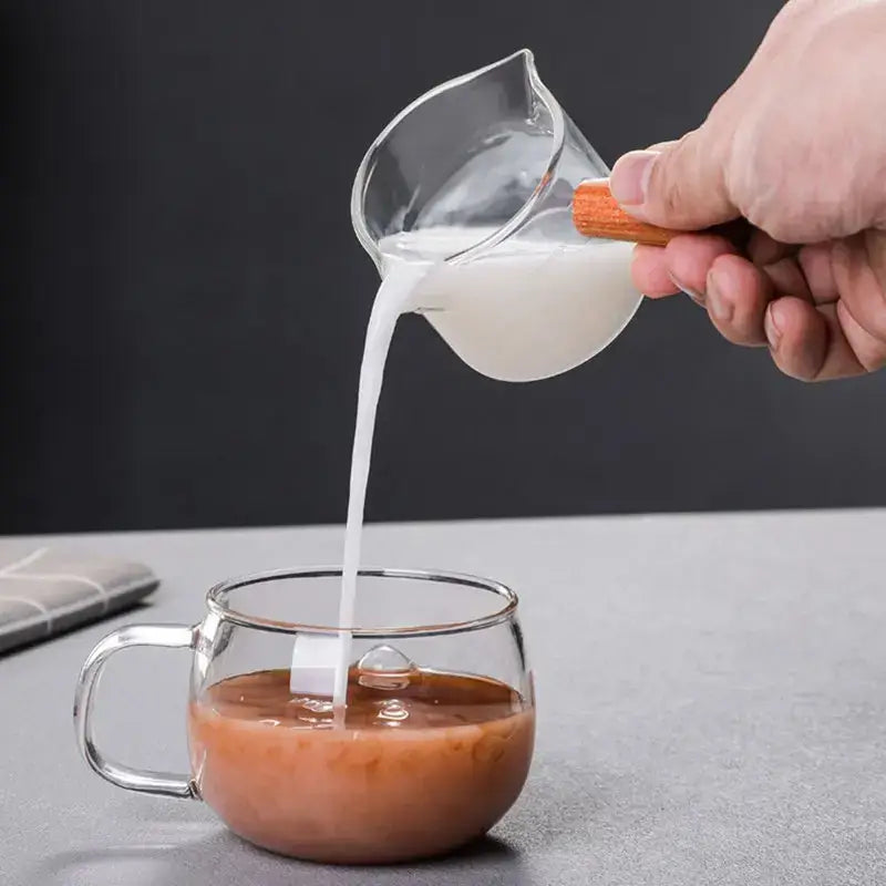 Heat resistant clear glass espresso milk jug with wooden handle pouring milk into matching glass mug