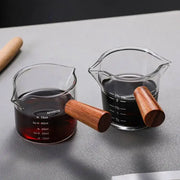 Two clear heat resistant glass espresso milk jugs with wooden handles filled with dark liquid and measurement indicators