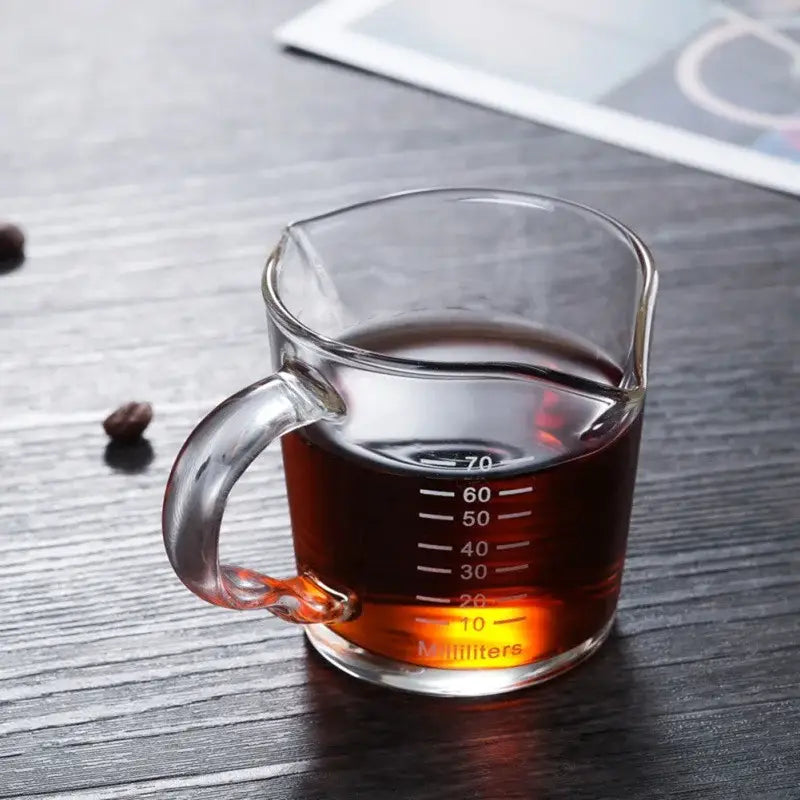 Heat resistant glass espresso milk jug with wood handle filled with dark liquid in a clear glass measuring cup