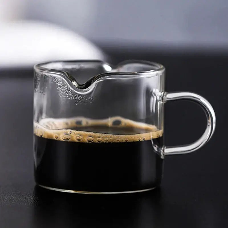 Heat resistant clear glass espresso milk jug with wood handle filled with dark espresso topped by golden crema