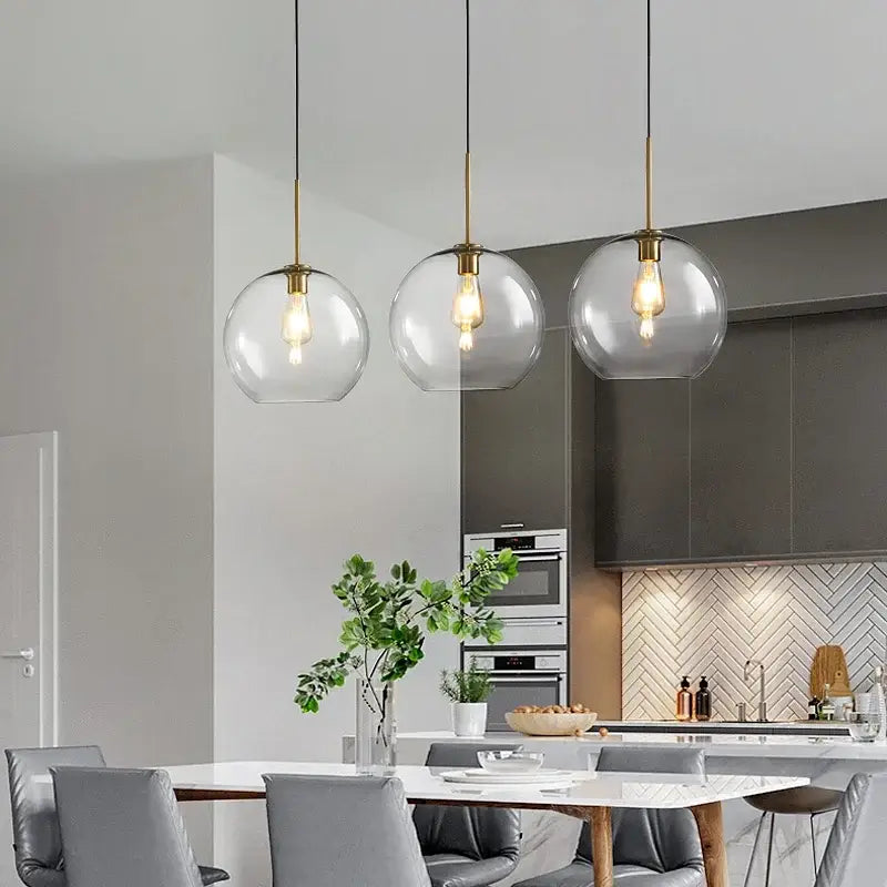 Three modern brass pendant lights with clear glass globes hang above a dining table in decorbites industrial loft glass style