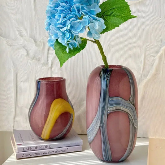 A tall blush-pink irregular geometric graffiti glass vase with blue and gray patterns holds bright blue hydrangeas