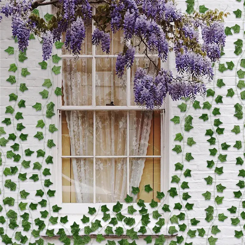 Purple wisteria blossoms hang from a garland vine with ivy green fake leaves on a white brick wall outdoor decor