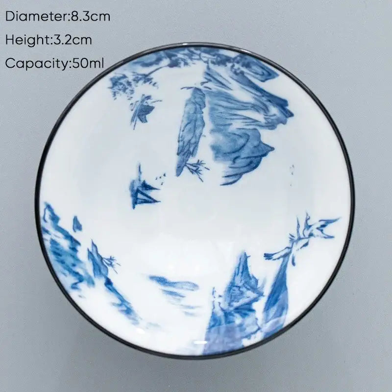 A white ceramic kung fu tea cup with black rim and blue hand-painted mountain and bird landscapes