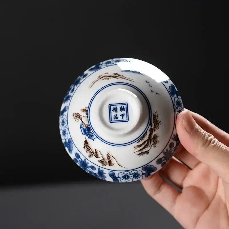 A delicate white ceramic kung fu tea bowl with blue and brown hand-painted landscapes and a central seal mark