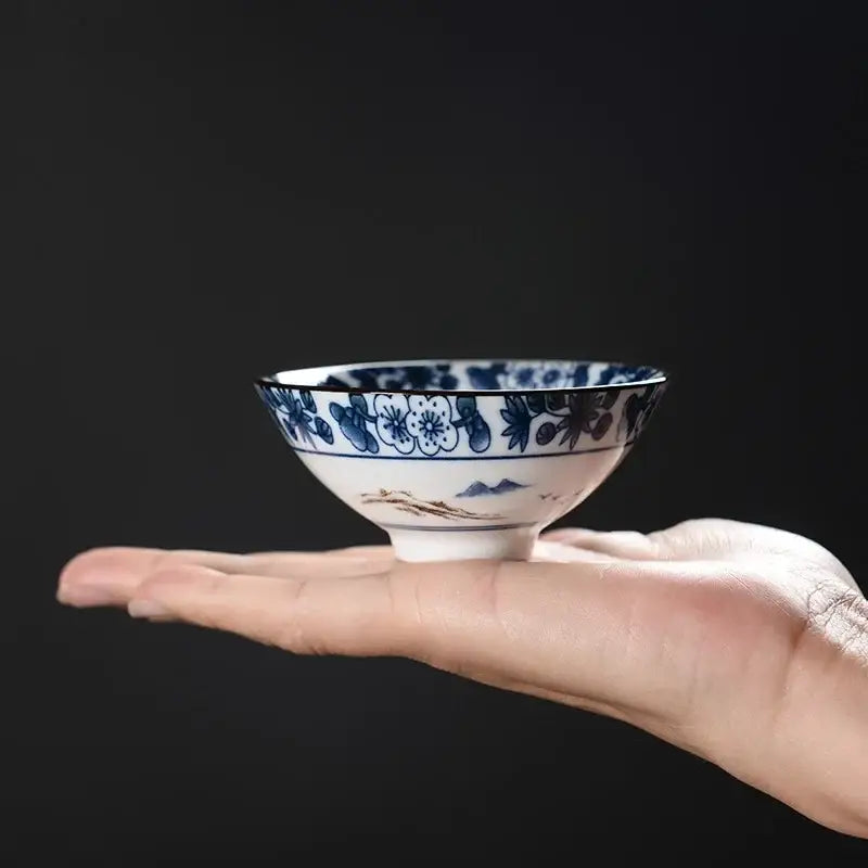 A delicate white ceramic kung fu tea cup with intricate blue floral and landscape motifs and brown base accent decorbites blue