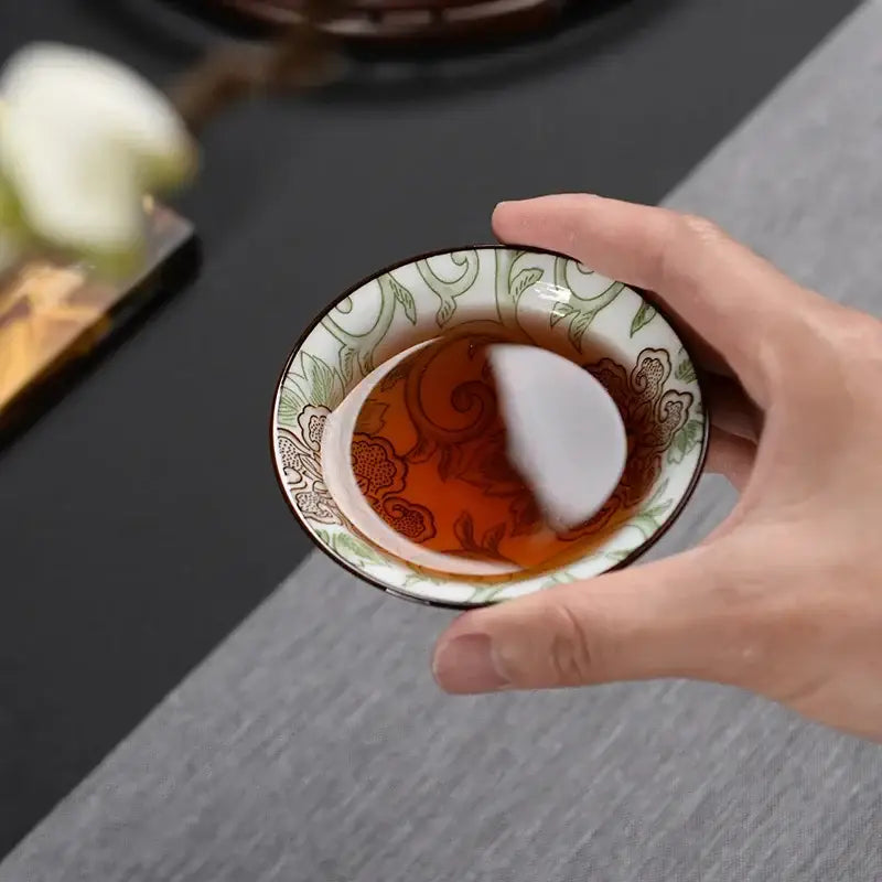 A hand holds a white ceramic kung fu tea cup with green brown floral patterns from decorbites blue set