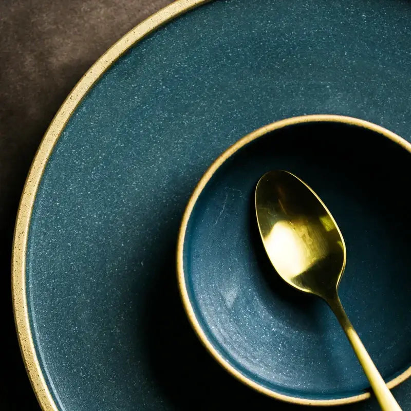 A matte teal decorbites ceramic dinner plate with gold rim paired with matching bowl and golden spoon for elegant dining