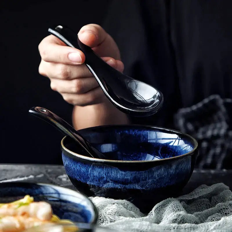 A glossy black ceramic spoon held above a kiln glazed blue speckled ceramic bowl for dining room ramen by decorbites kiln glazed