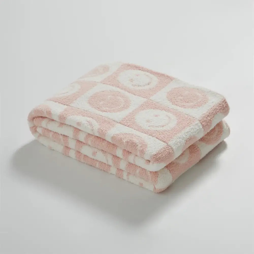 A soft pink and white knitted checkboard smile face blanket with plush embroidered smiley faces decorbites decor