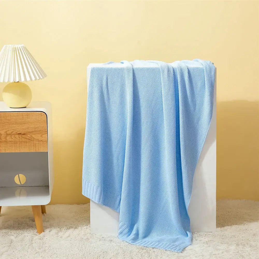 A soft light blue knitted bamboo baby blanket with textured weave draped over a white pedestal for nursery use