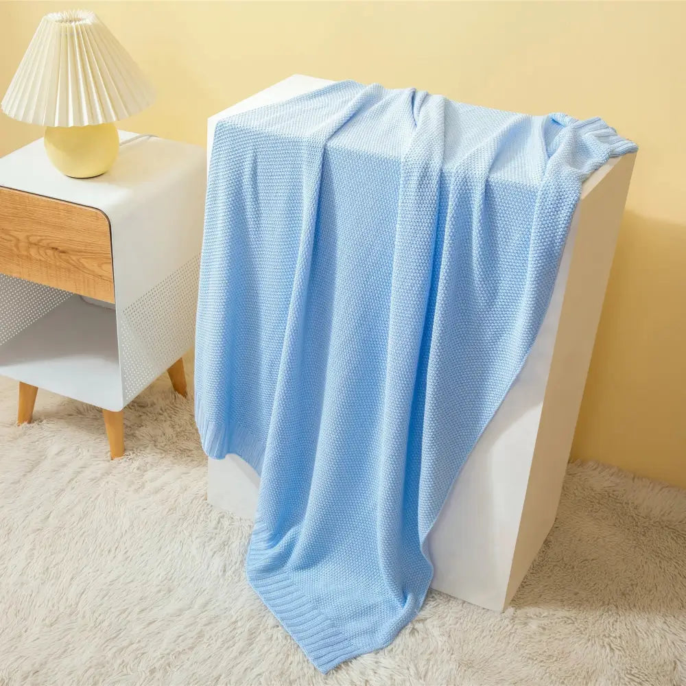 A soft light blue knitted bamboo baby blanket with textured weave draped over a white cube for nursery use