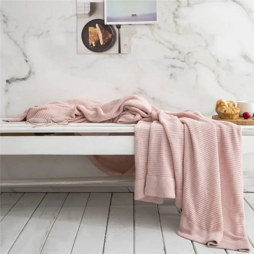 A soft pink knitted bamboo cooling throw blanket with waffle weave pattern draped over a white wooden bench