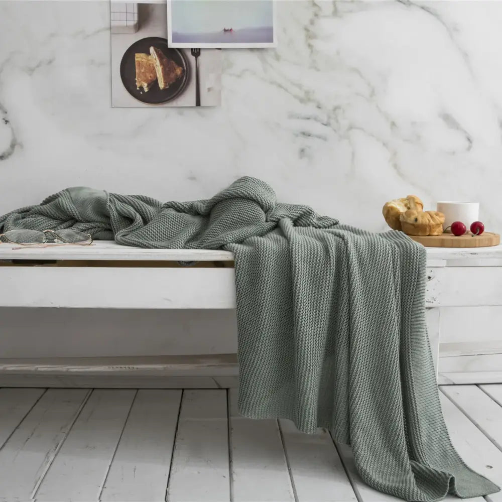 A soft sage green knitted bamboo cooling throw blanket with textured weave draped over a white wooden bench