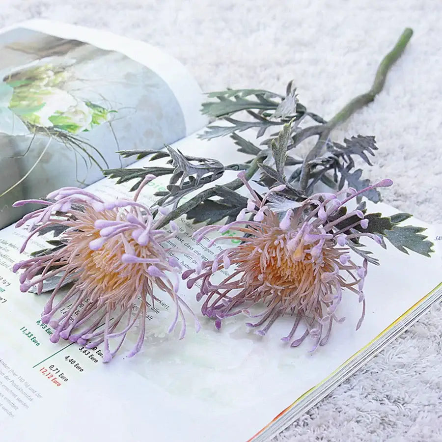 Two artificial purple and peach chrysanthemum flowers with detailed petals and green stems on an open book for decorbites artificial crab claw branch decor