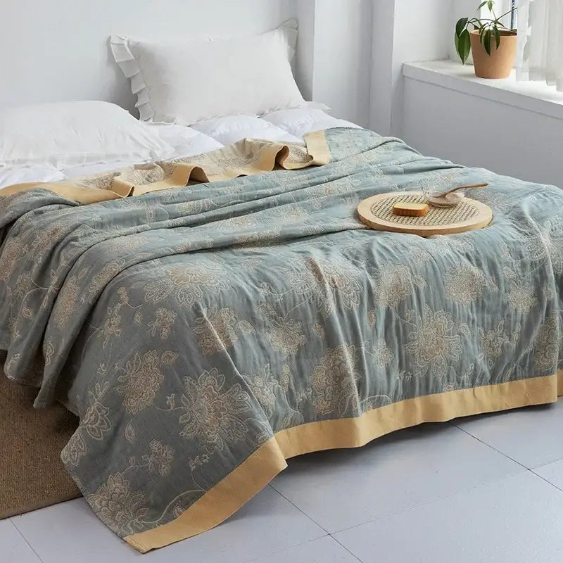A soft sage green quilted bedspread with gold floral embroidery and mustard border contrasting the decorbites cotton gauze towel throw