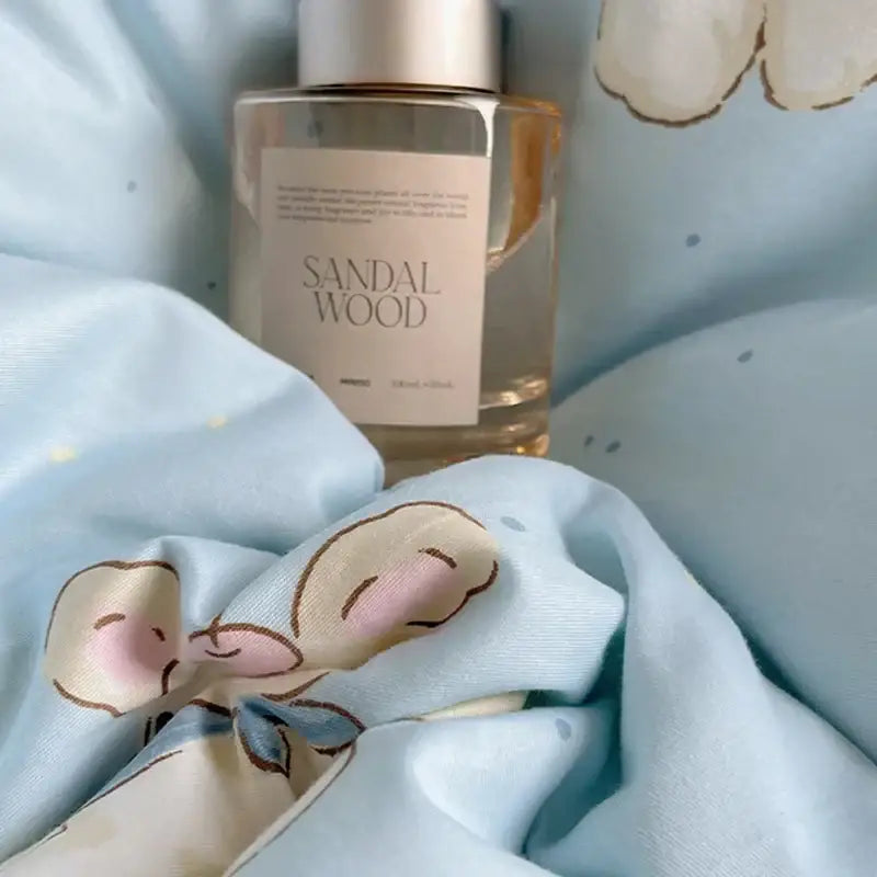 A clear glass bottle with amber sandalwood fragrance next to a light blue puppy cartoon pattern bedding set cotton sheets
