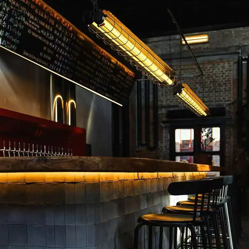 Industrial-style linear chandelier glass pendant lamp with warm yellow glow and metal wire cages hanging above a bar