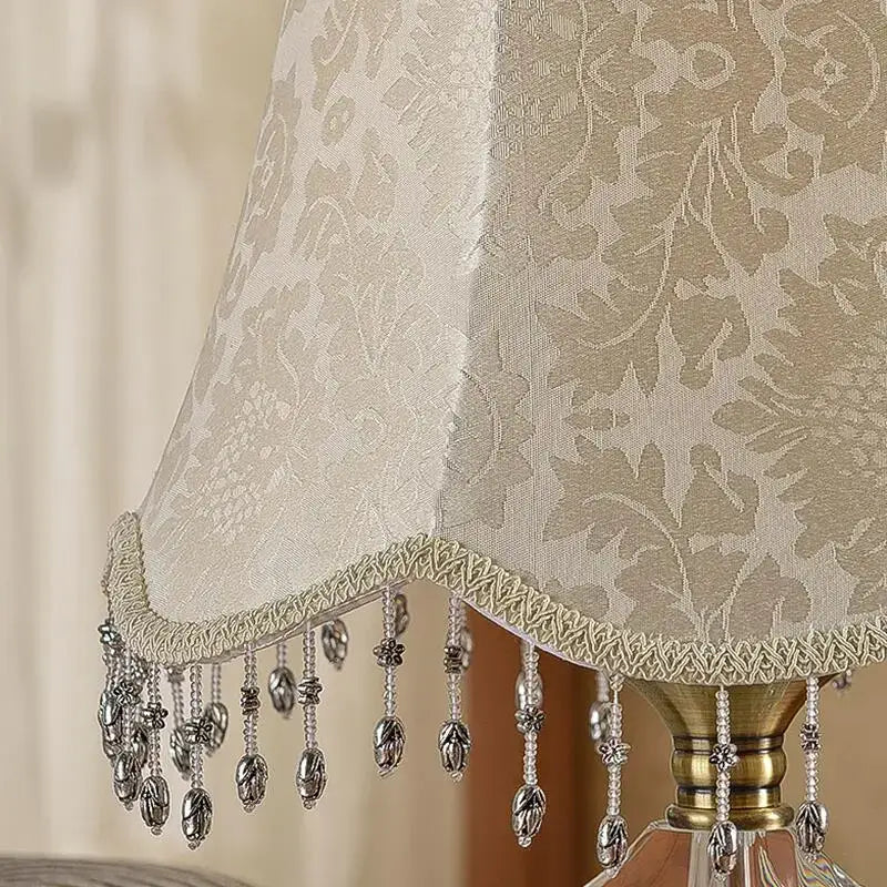 Luxury crystal table lamp with beige fabric lampshade featuring silver beaded fringe and metallic leaf-shaped tassels by decorbites